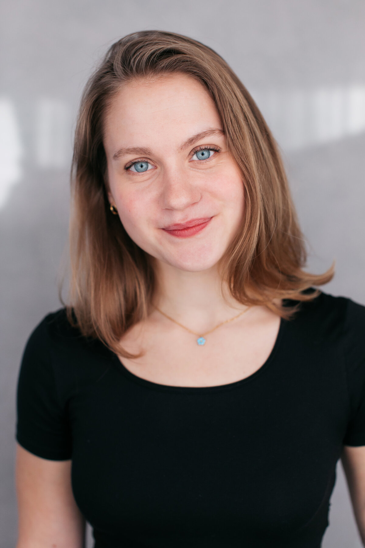 Castingfoto van jonge vrouw met schouderlengte donkerblond haar in zwart t-shirt voor grijze achtergrond.