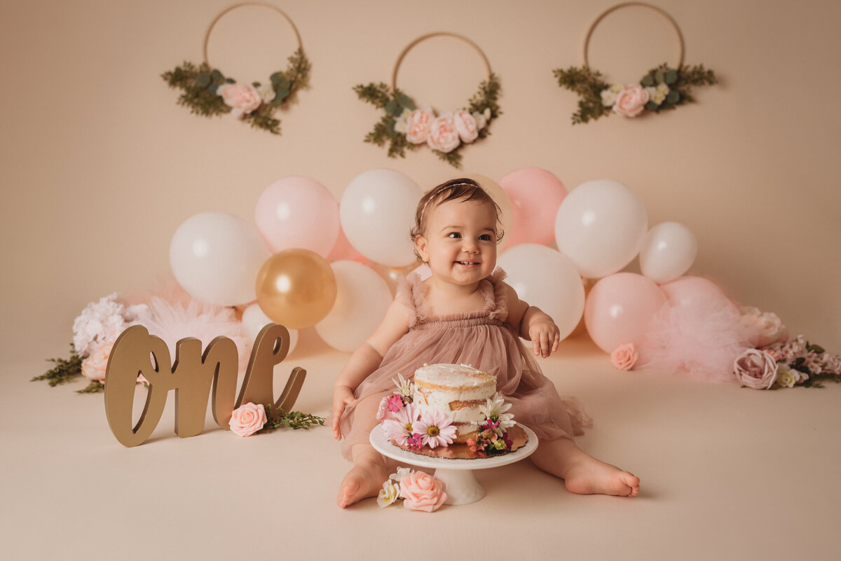 One year milestone cake smash at Marietta GA portrait studio with baby girl wearing pink tulle dress sitting on floor eating cake decorated with flowers. Balloons are behind her with a gold O N E sign and floral decor on the tan backdrop