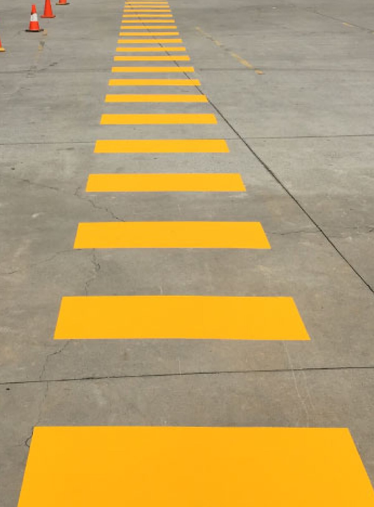 A painted yellow line marking for pedestrian walkways on a concrete carpark with orange safety cones.