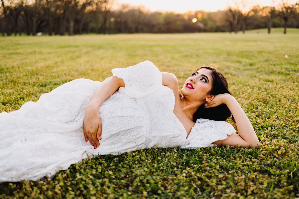 Photo of a pregnant woman lying on the grass in a garden, her head is on her arm and she is wearing a white dress and is looking up