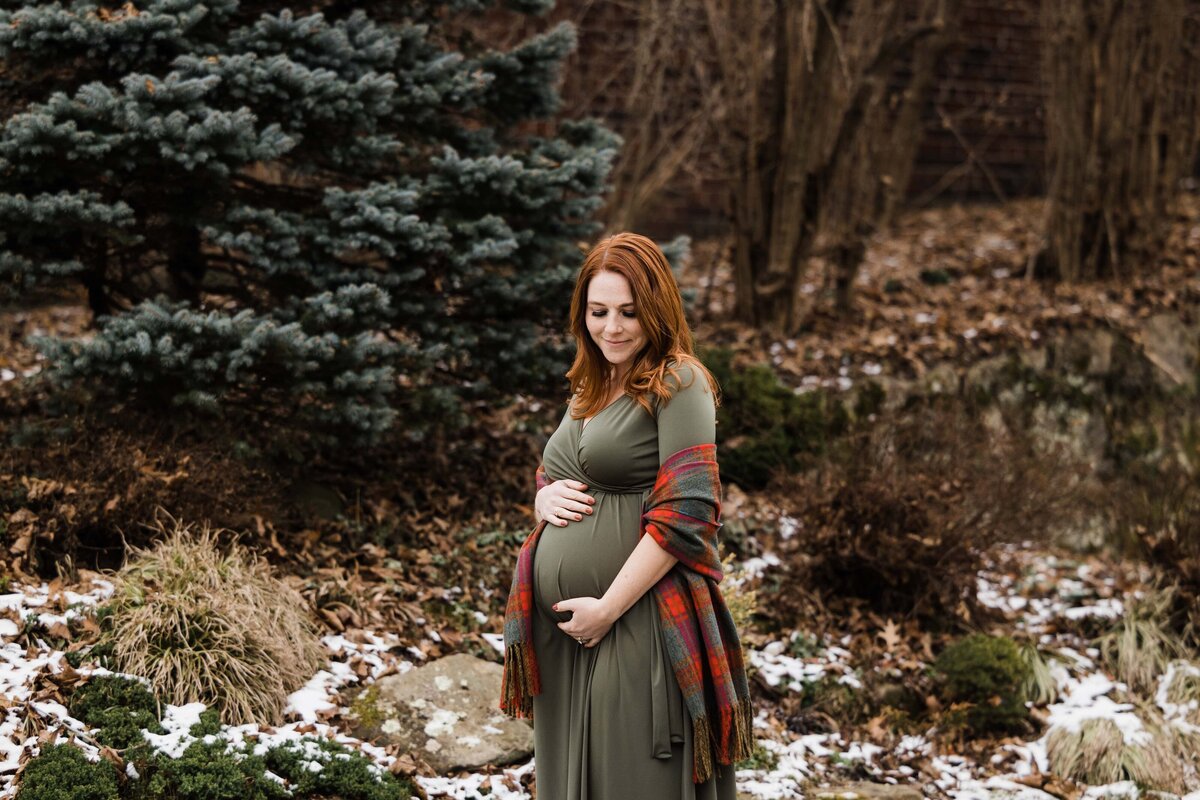 A pregnant woman holding her belly stands outdoors near evergreen trees with patches of snow on the ground, creating a perfect moment for maternity photography.