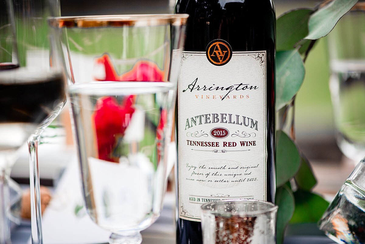 Bottle of Arrington Vineyards Antebellum red wine sitting on a farm table at a wedding next to a footed crystal water goblet with a gold rim.