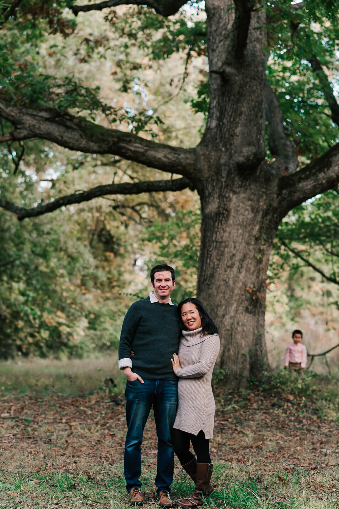 A couple standing together in front of a tree while their son stands in the distance