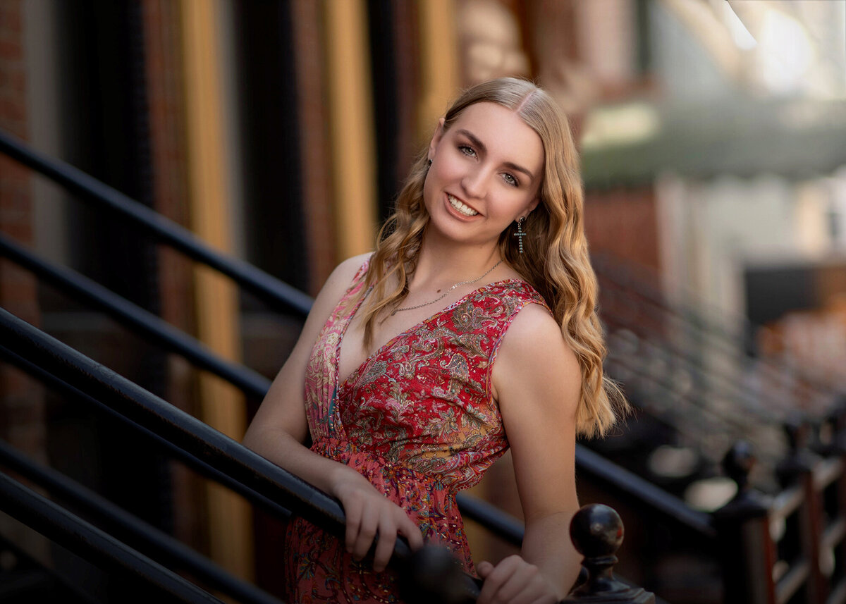 An image from a Pella Senior Photographer. A blonde high school senior in a red print dress walks up stoop steps holding the metal railing