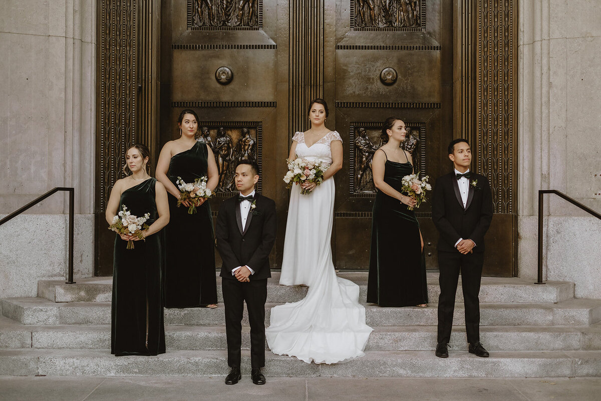 A wedding party stands in front of the wooden door to Theatre St James, holding bouquets.