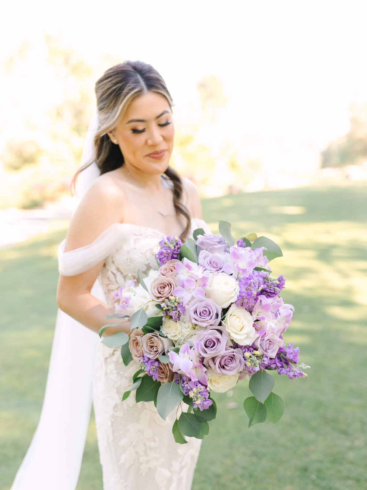 Bride holding a bouquet of pastel roses and lilacs. She wears an off-shoulder lace gown, smiling softly, with a sunlit garden in the background.