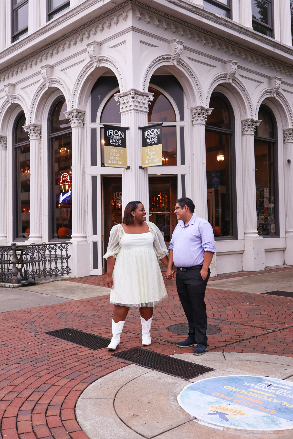 woman wearing white short tulle dress and white cowgirl boots and man wearing black dress pants and purple shirt holding hands looking at each other in front of gray stone building