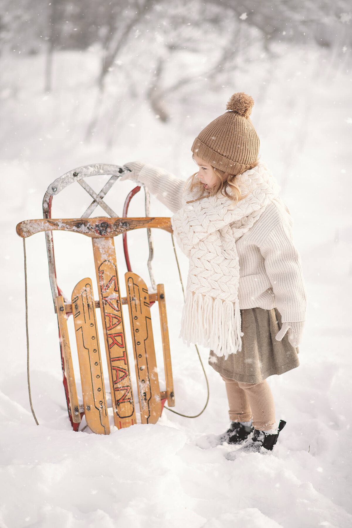 Toddler girl wearing a brown toque holds up a wooden sled in the snow, admiring it.  Portrait by Brittany Danielle.