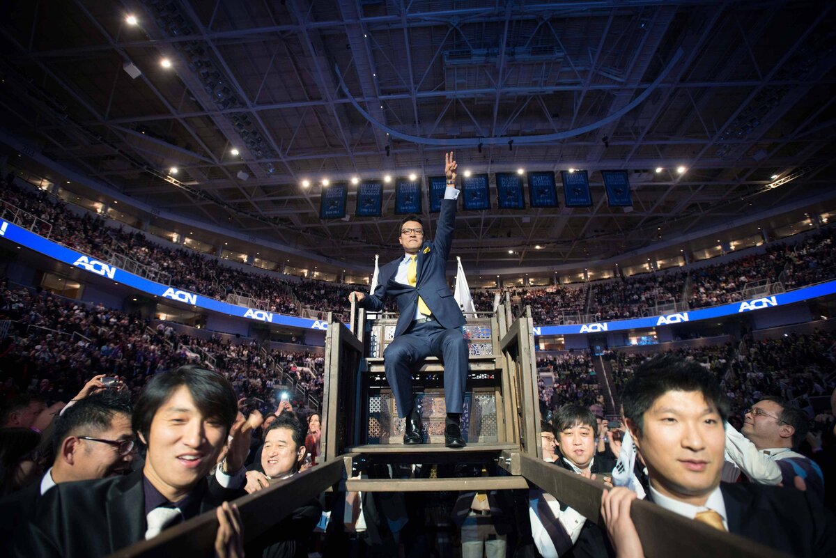 A man is carried on a chariot  at  a networking marketing event surrounded by cheering crowd
