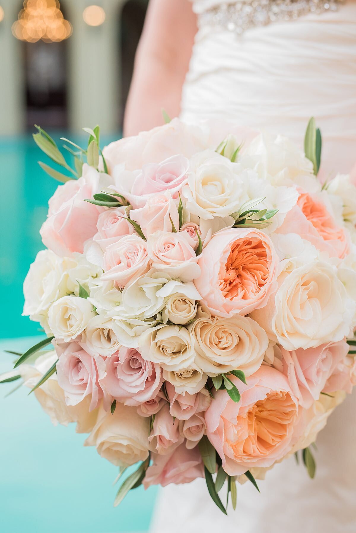 A bride in a fitted wedding dress holds a large round bouquet of peach peonies, ivory tea roses, blush tea roses, light pink roses, ivory roses and greenery.