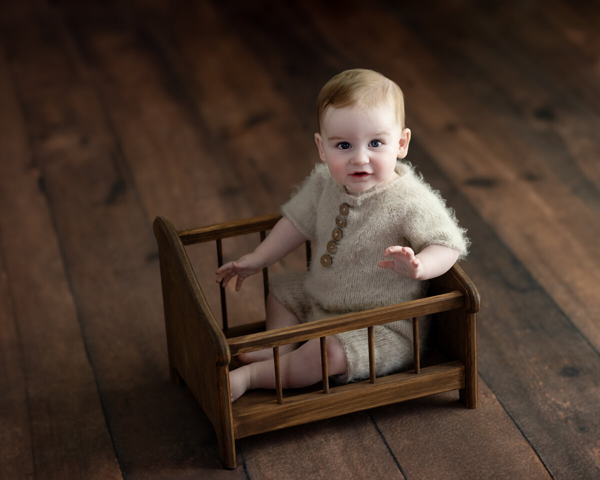  Rustic studio portrait of baby boy sitting in a wooden bed wearing a cozy knit romper—timeless textures and charm.