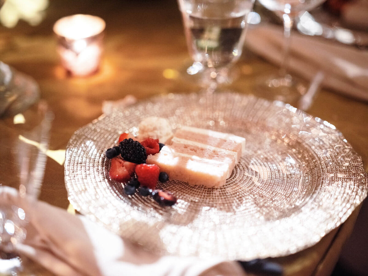 A small slice of a pink cake with mixed berries on the side, on a glass plate, with glasses of water and a candle in the background