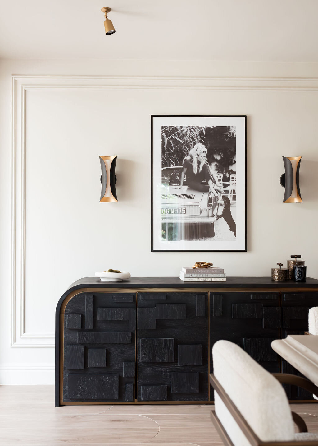 A modern dining room features a large light-colored table with beige padded chairs. On the wall, a black sideboard with a photograph above it is flanked by two contemporary sconces. The space is minimalistic with neutral tones and a sculptural white centerpiece on the table.