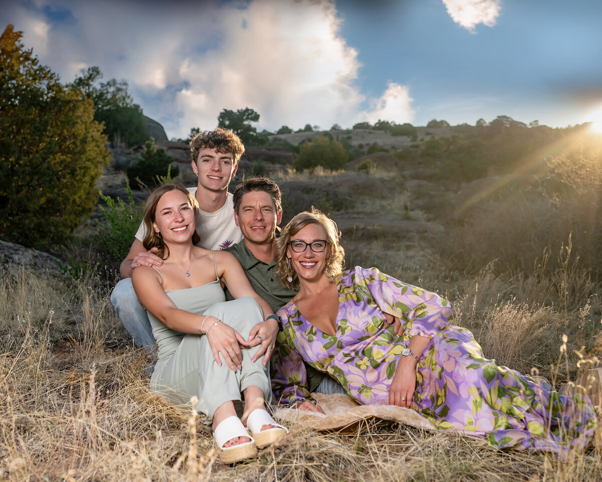 A family of four is smiling and relaxing together in a dry, grassy field under a partly cloudy sky with the sun setting behind them.