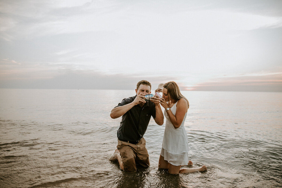 Fun engagement photo of man and woman kneeling in the shores of Lake Huron in Grand Bend shotgunning a beer at golden hour.