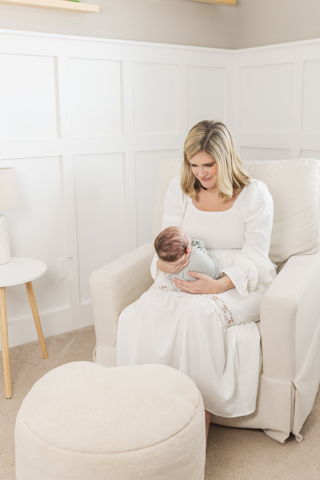 Mom sits in glider in nursery looking down at her swaddled newborn in her arms.