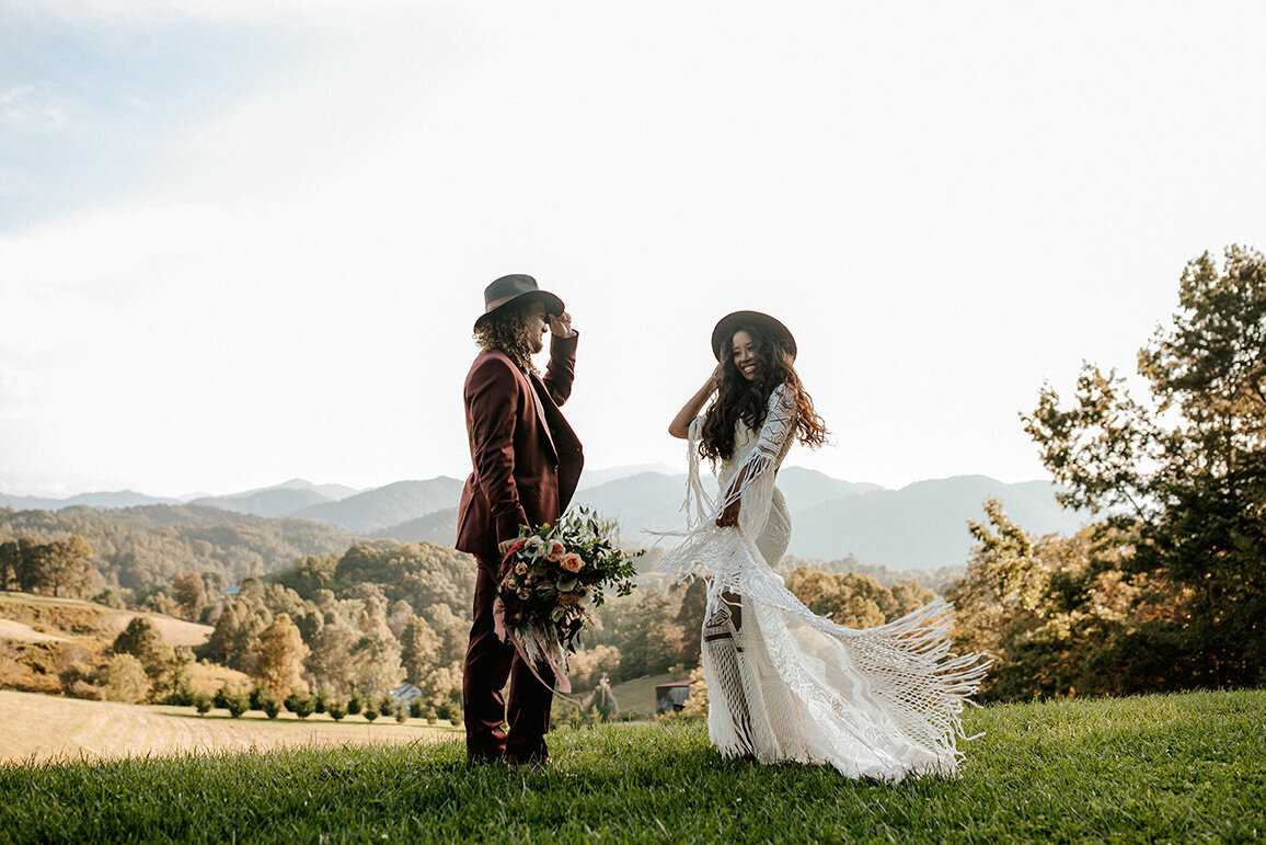 bride and groom wearing a white wedding gown and burgundy tuxedo  with hats posing against a mountain view