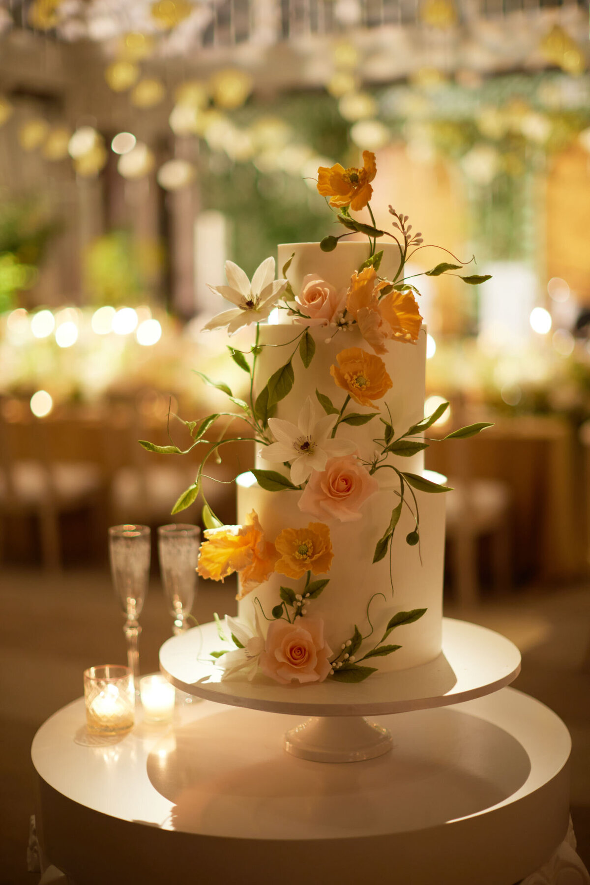 This stunning three-tiered white wedding cake adorned with yellow and peach flowers, greenery, and delicate vines was captured at the exquisite New York City location, The New York Public Library, by the talented photographer Shawn Connell. Videography was beautifully done by Aaron Navak Films. The cake is elegantly displayed on a white pedestal, complemented by two elegant champagne flutes and a softly glowing candle nearby. The background showcases a beautifully decorated reception area with twinkling lights, adding to the enchanting ambiance of the occasion.