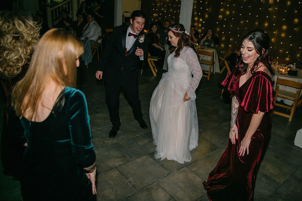 A bride in a white dress dances with a groom in a black suit and two women in velvet dresses, surrounded by guests seated at tables and warm string lights in the background.