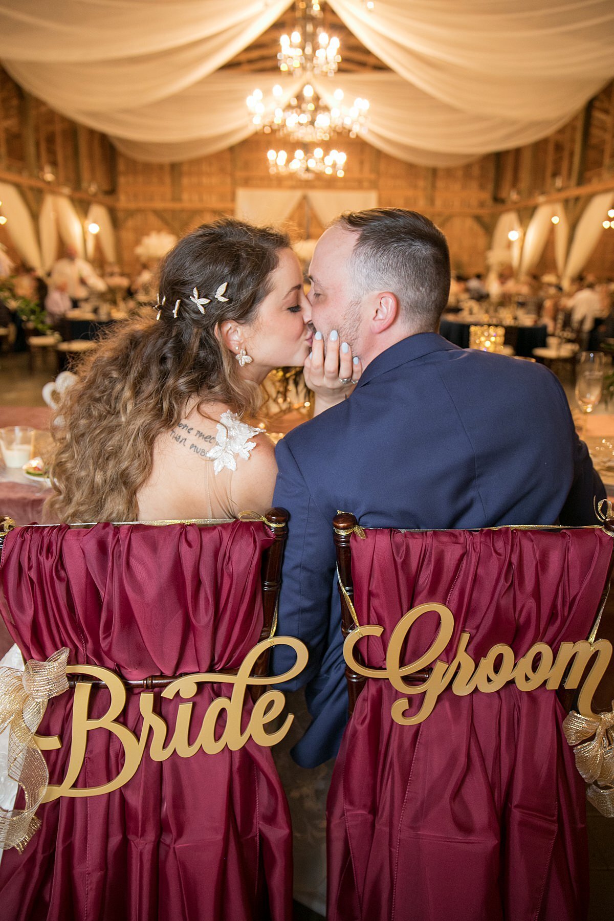 The bride and groom kiss at their Saddle Woods Wedding reception while sitting in their specially decorated chiavari chairs covered with burgundy organza fabric and gold BRIDE and GROOM calligraphy signs. The ceiling is draped in sheer ivory fabric accented by crystal and gold candelabra chandeliers.