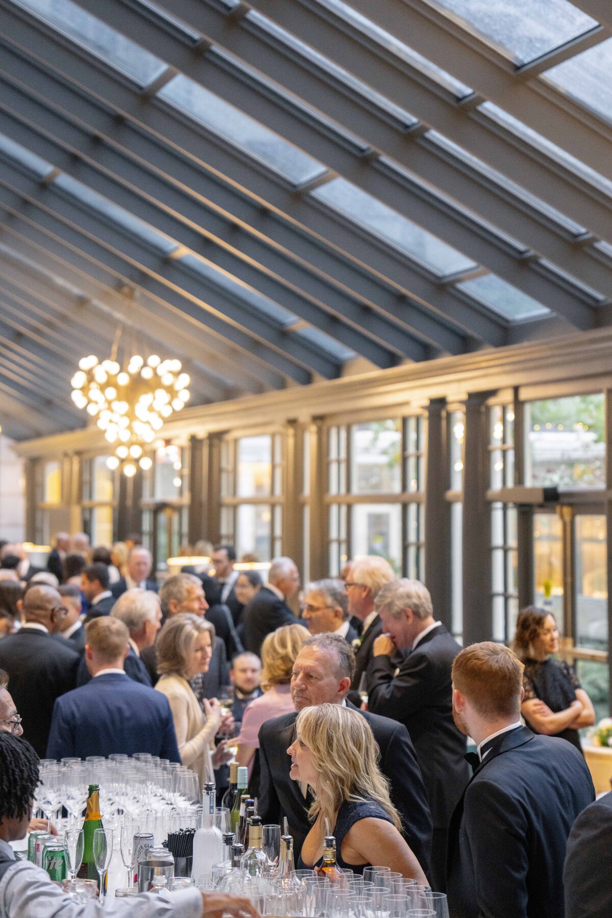 A crowded indoor gathering with people mingling under a glass ceiling. A table with drinks and glasses is in the foreground. Soft lighting creates a warm atmosphere, and a decorative chandelier hangs overhead.