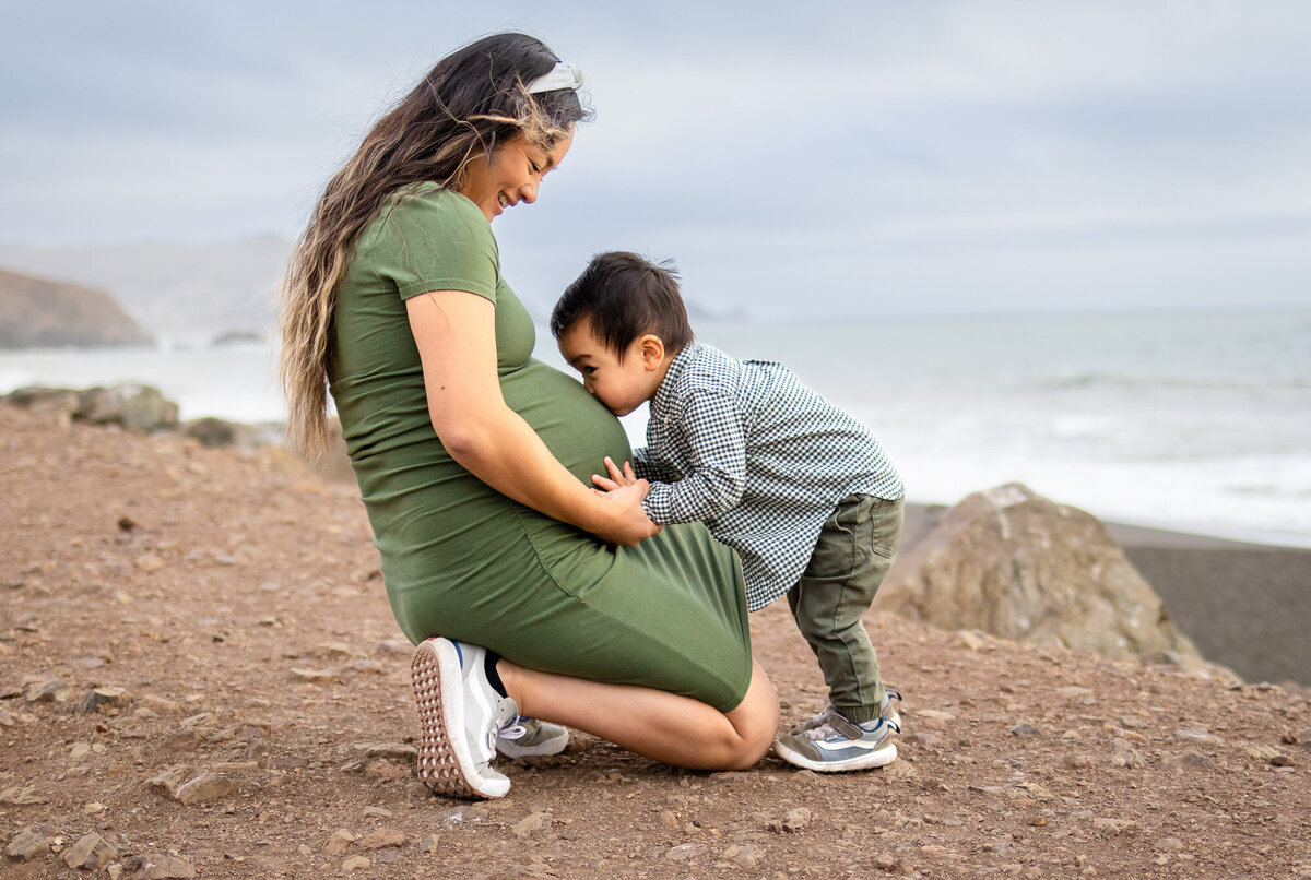 Toddler kissing mom’s pregnant belly while kneeling on a dirt path by the ocean, Bay Area Maternity Portfolio - Ellobelle Photography