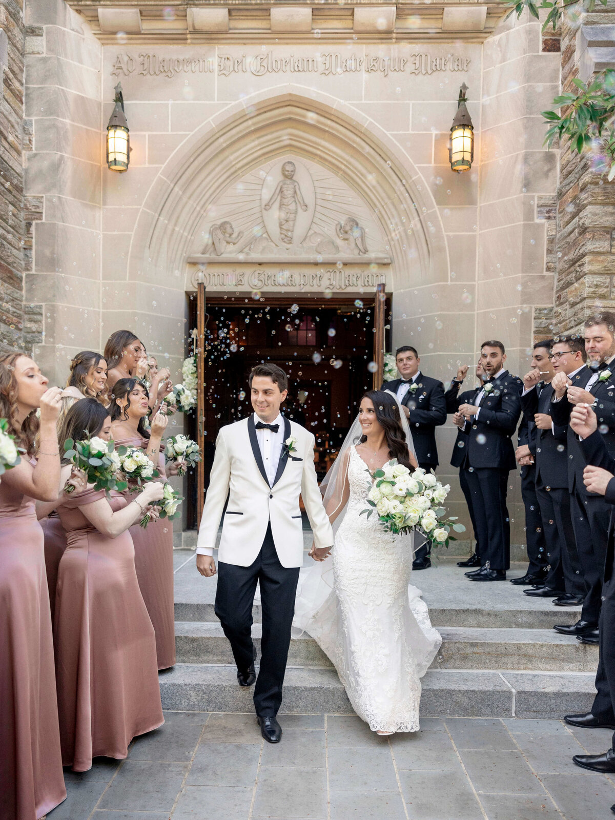 A newlywed couple exits a church, smiling, surrounded by a wedding party. The bride wears a white gown and holds a bouquet, while the groom wears a white tuxedo jacket. Bridesmaids and groomsmen stand nearby, dressed in formal attire, celebrating with bubbles.