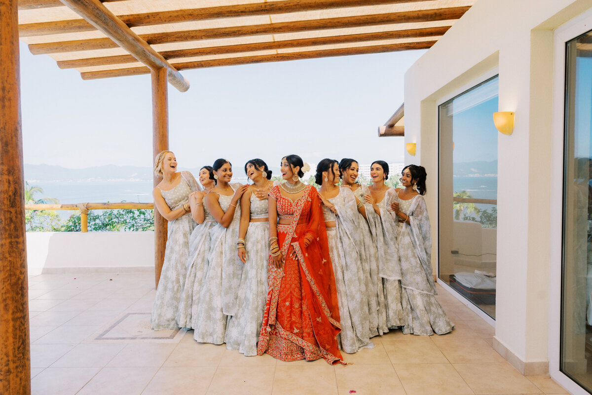 A bride in a red lehenga stands in the center of her bridesmaids, who are wearing light gray patterned dresses. They are standing on a terrace with an ocean view in the background, posing for a wedding photo.