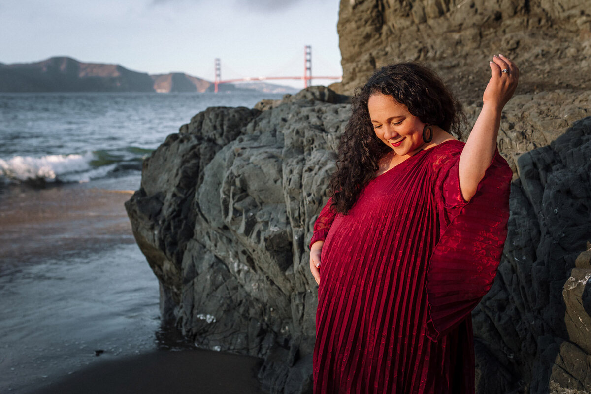 pregnant woman with arm raised and looking at her bump in front of a rock at China Beach in San Francisco with the golden  gate bridge in the background