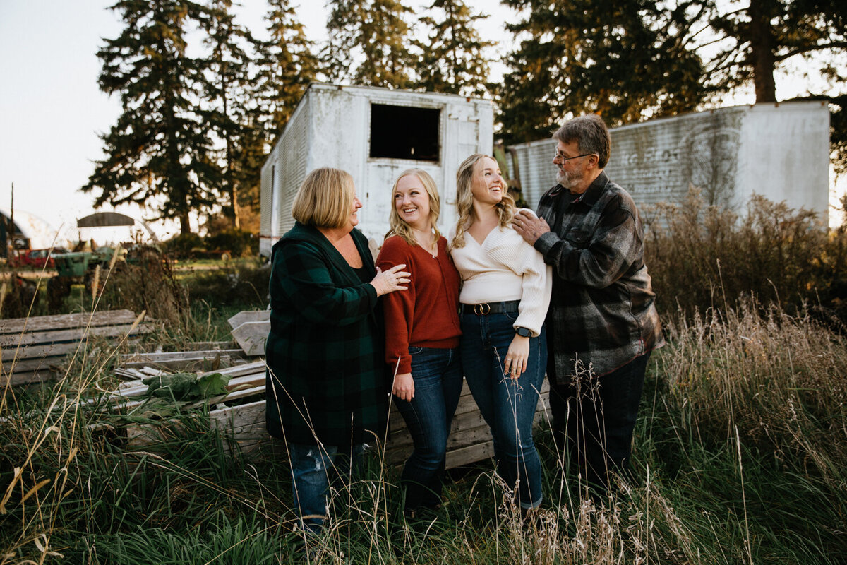 Parents and their adult children are standing in front of a rustic farm trailer. Mom and dad have their hands on their adult children's shoulders They are all looking at each other and smiling.