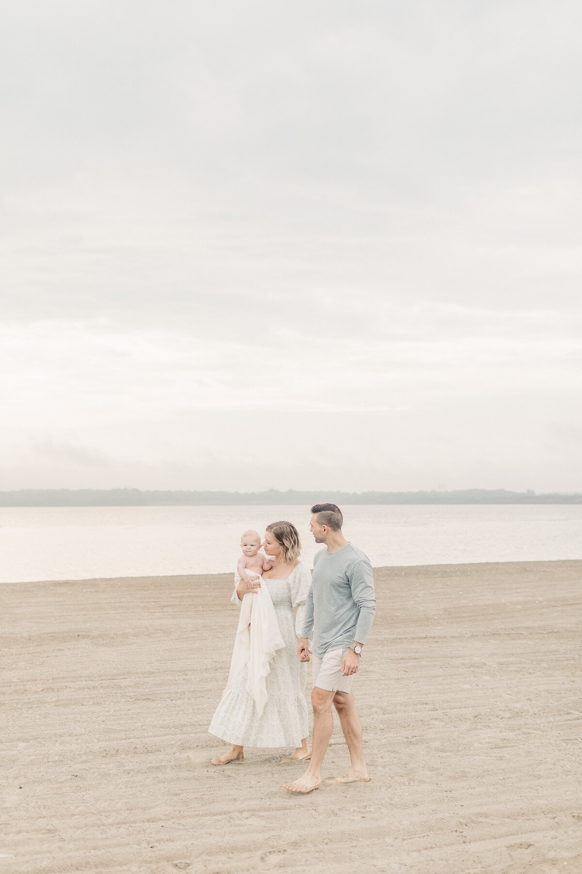 Mom and dad walking together with their baby in mom's arms along the shoreline during a Kansas City lake milestone session.