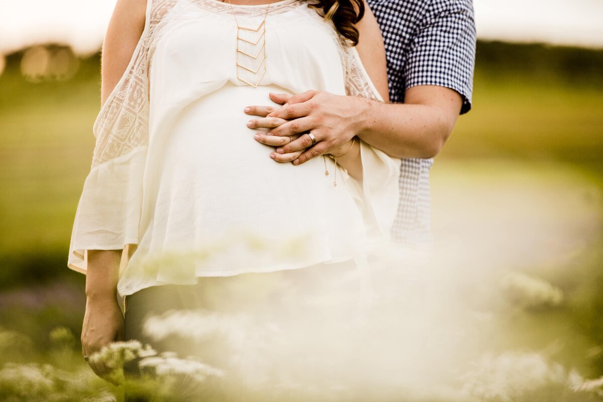 A couple standing close together, with the man's hands around the woman's pregnant belly in a grassy field, captured by a Pittsburgh maternity photographer.