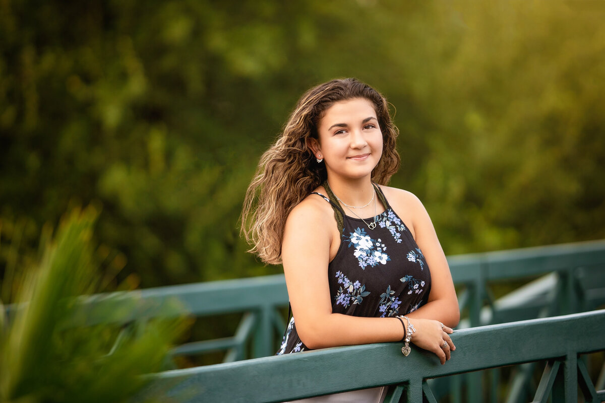 Senior girl in San Antonio on a bridge.  She has dark curly hair and is leaning on the rail of a pedestrian bridge.  The bridge is painted green.  She is wearing a black floral top.  There are lots of trees around her.