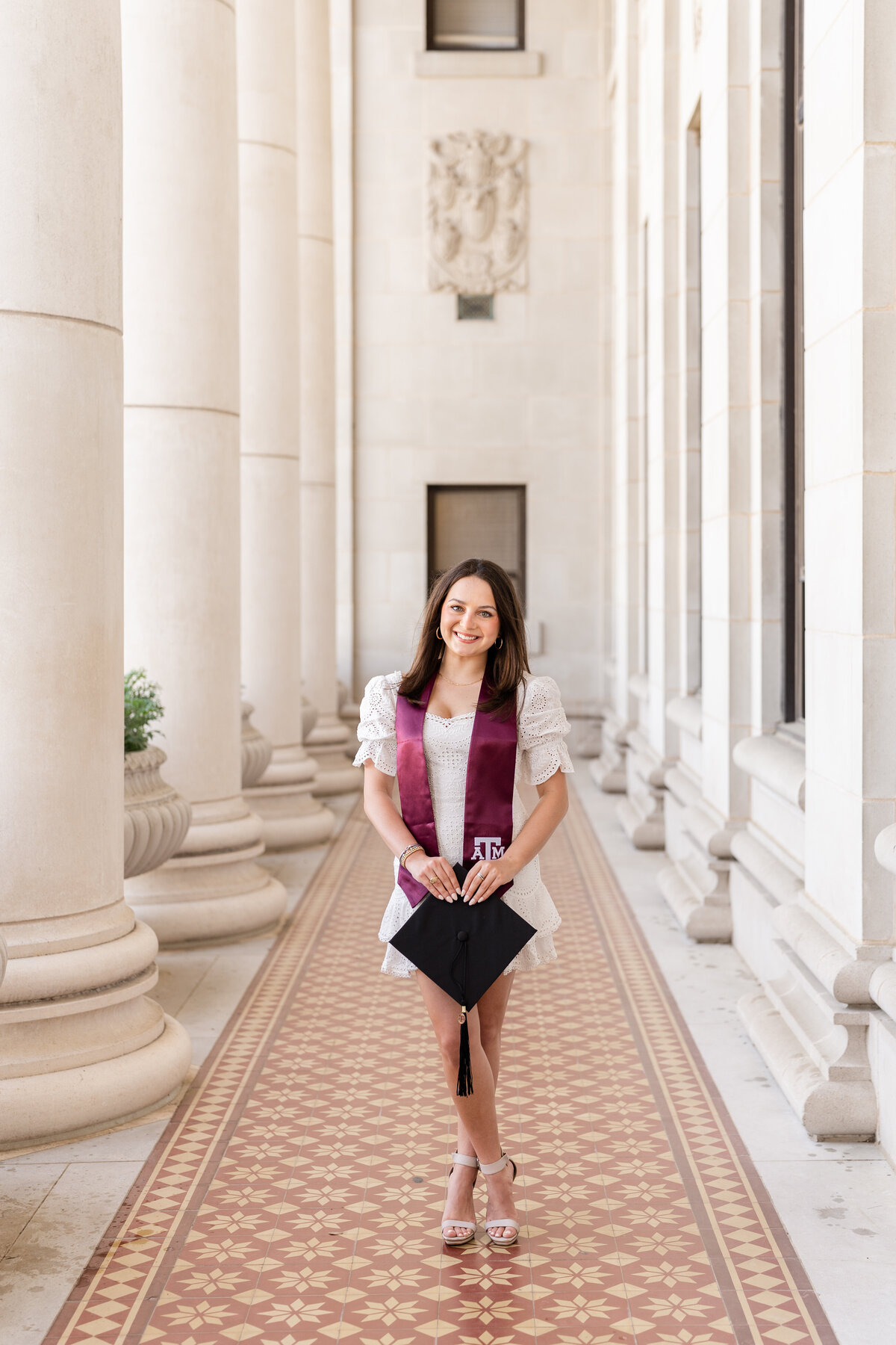 Texas A&M senior girl standing and holding grad cap while wearing white dress and Aggie stole  within columns of the Administration Building