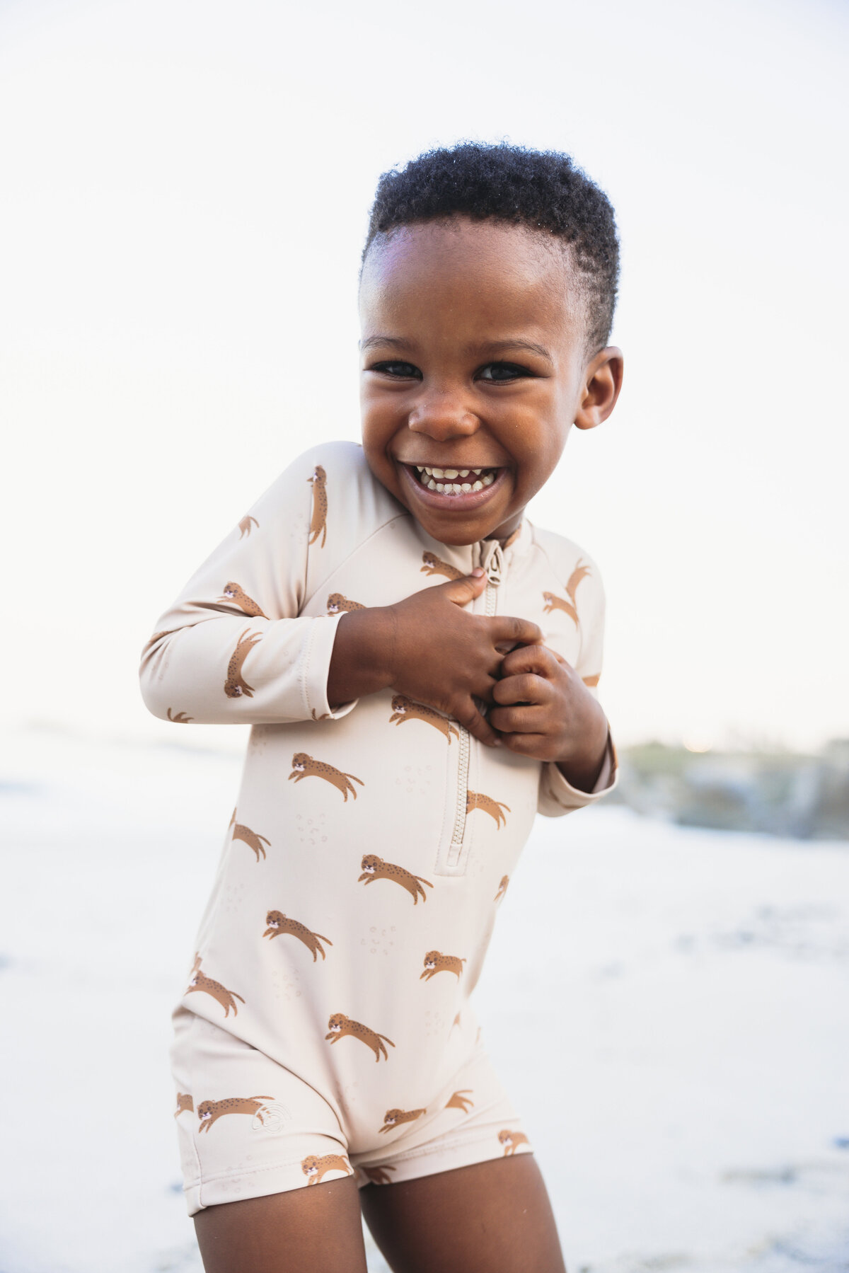 Smiling young boy wearing a beige UV swimsuit with cheetah print, standing on a bright sandy beach in Cape Town.