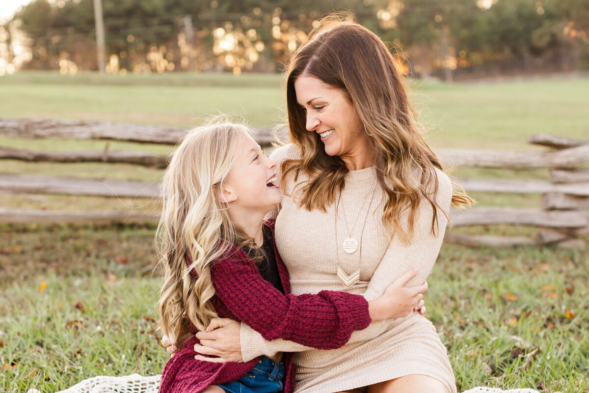 Mom and daughter sit on blanket in a field and hug each other while looking at each other