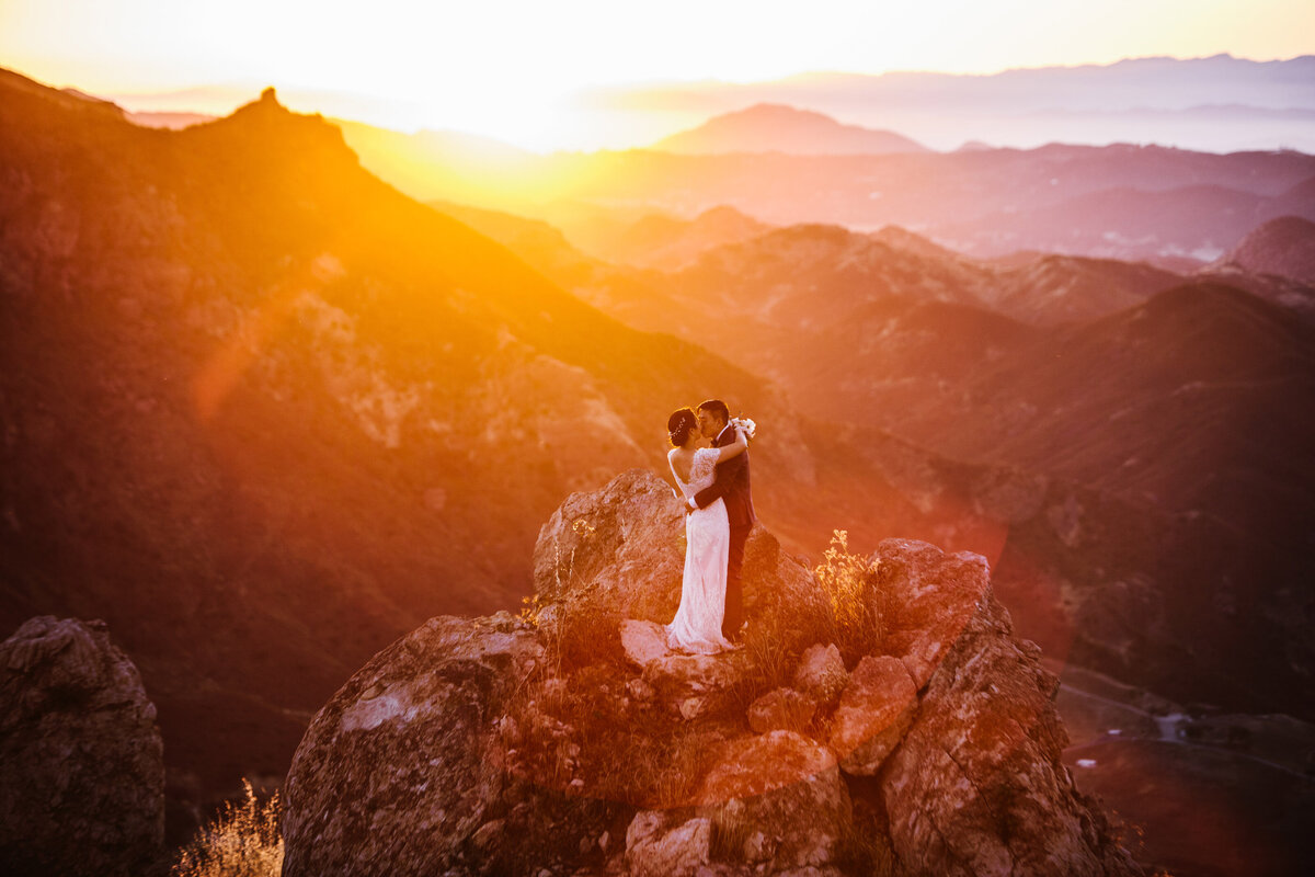 bride and groom kiss on a mountain top with a colorful sunset behind them