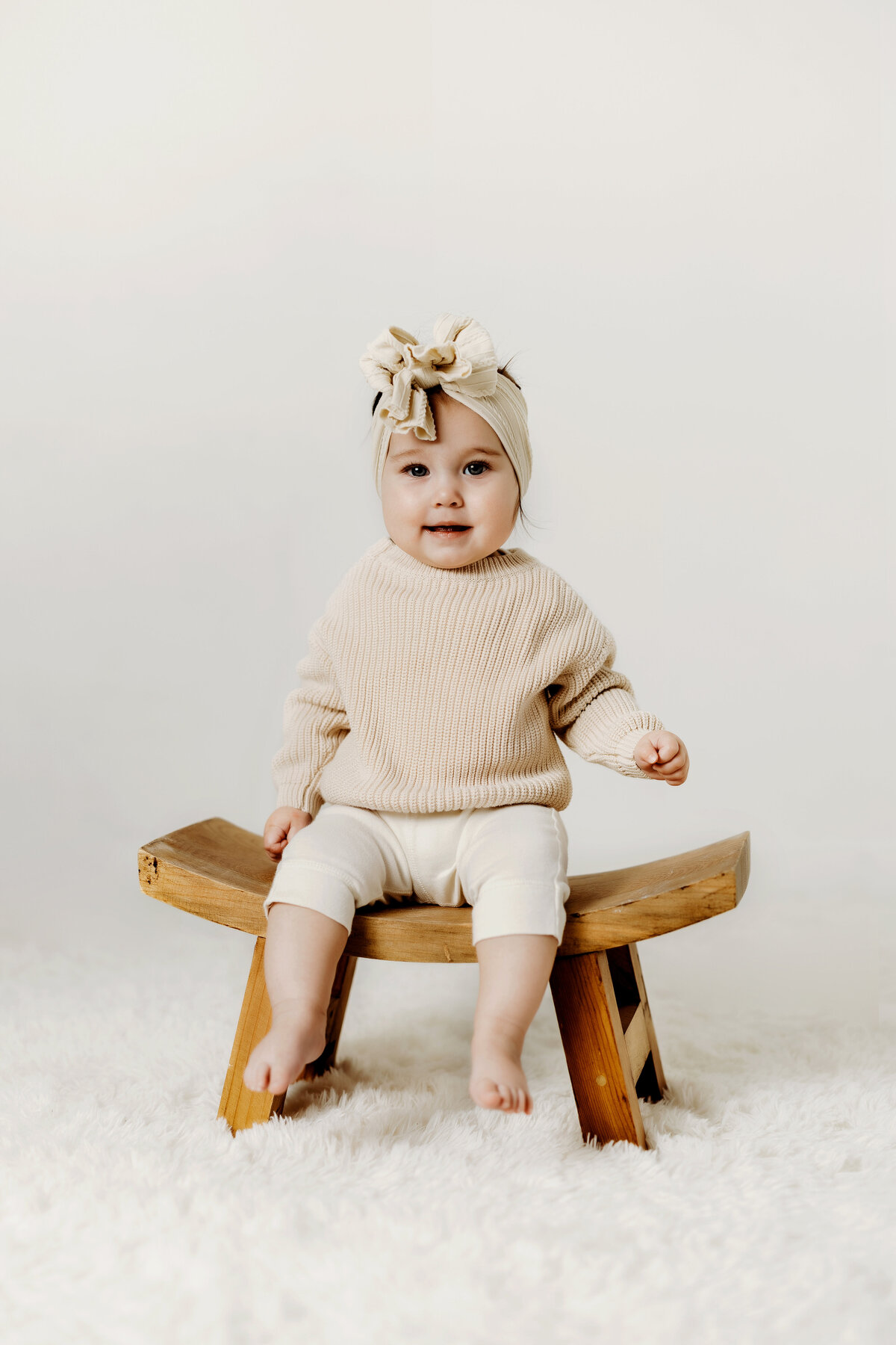 A happy eight month old girl sits on a wooden chair on a white rug while wearing a knit sweater during her milestone session in Denver. 