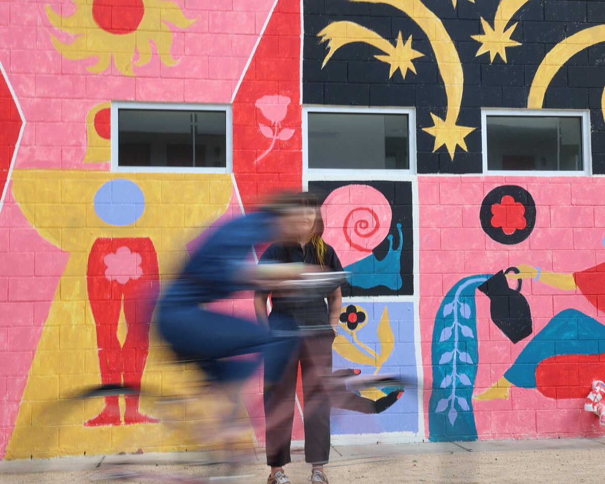Blurred image of woman in a bike riding past another person with a colorful mural in the background