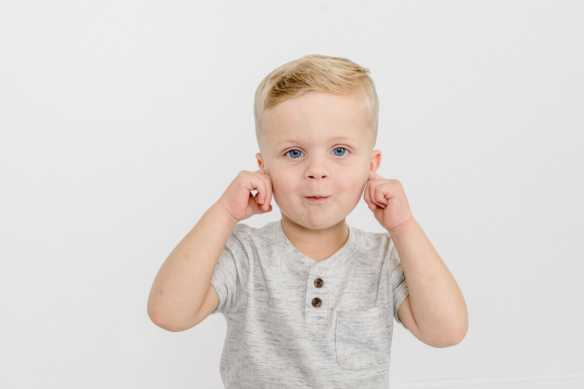 Close-up of a little boy with blonde hair in a grey shirt with his hands on his cheeks, pulling his cheeks out in a silly face and looking at the camera in front of a white background.