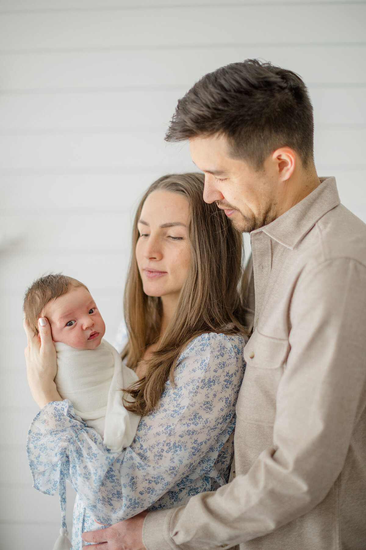 In a cozy in-home lifestyle newborn session, a mother cradles her newborn in her arms while she and the father gaze lovingly at their baby. Soft natural light fills the room, highlighting the warmth and intimacy of the moment. The parents' expressions reflect pure love and joy as they soak in the precious early days with their little one.