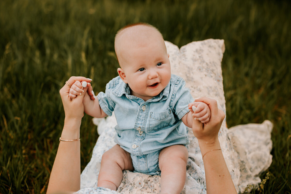 six month old baby boy sits on moms thighs in a grassy field