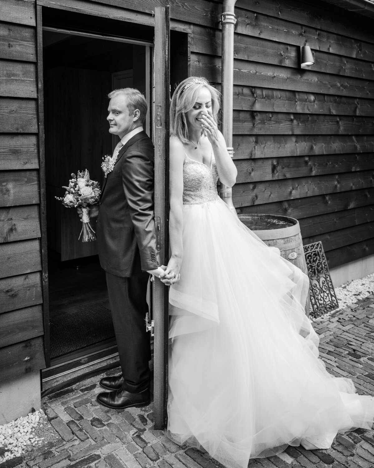 Black and white photo of a bride and groom standing back to back with a door separating them as they hold hands without seeing each other before the ceremony
