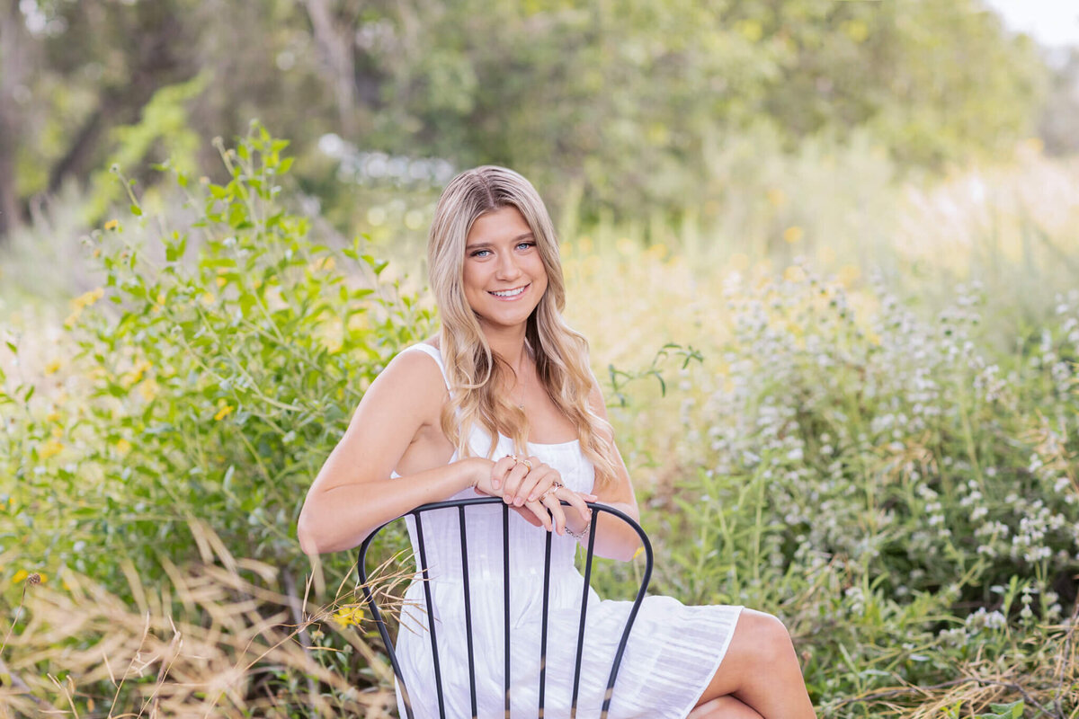 A person with long hair sits on a black chair in a lush, green garden. They wear a white dress and smile at the camera. The background is filled with plants and trees under soft, natural light.