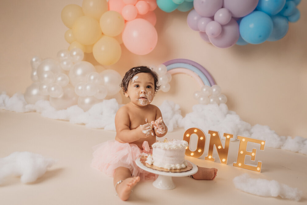Photo shoot at Atlanta, Georgia cake smash photographer studio with one year old little girl in pink tutu smashing a cake with rainbow pastel balloon garland behind her with white fluffy clouds.