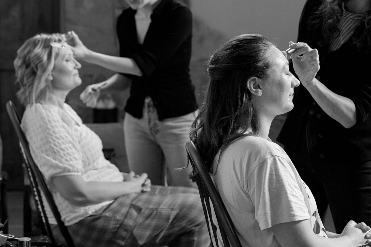 Behind-the-scenes moment captured at Willow Marsh Farm, where two women are getting ready for the wedding day. In this intimate black-and-white photo, one woman smiles as a makeup artist carefully applies makeup to her eyebrows. Another woman sits with her eyes closed, enjoying the pampering, as she gets her makeup done. The cozy, relaxed atmosphere of the farm provides the perfect setting for these special moments of preparation.