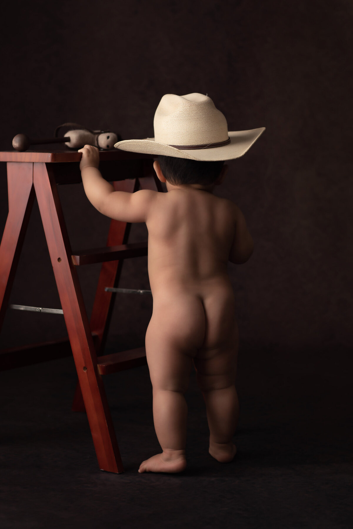 A playful baby wearing a cowboy hat stands confidently beside a small wooden ladder, showcasing curiosity and charm. This rustic-themed milestone photo captures the child's adventurous spirit, skillfully photographed by a Dallas baby milestone photographer.