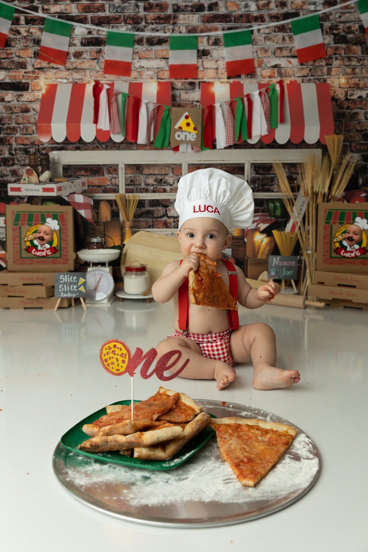 A toddler boy sits in a pizza themed studio eating a slice for his first birthday