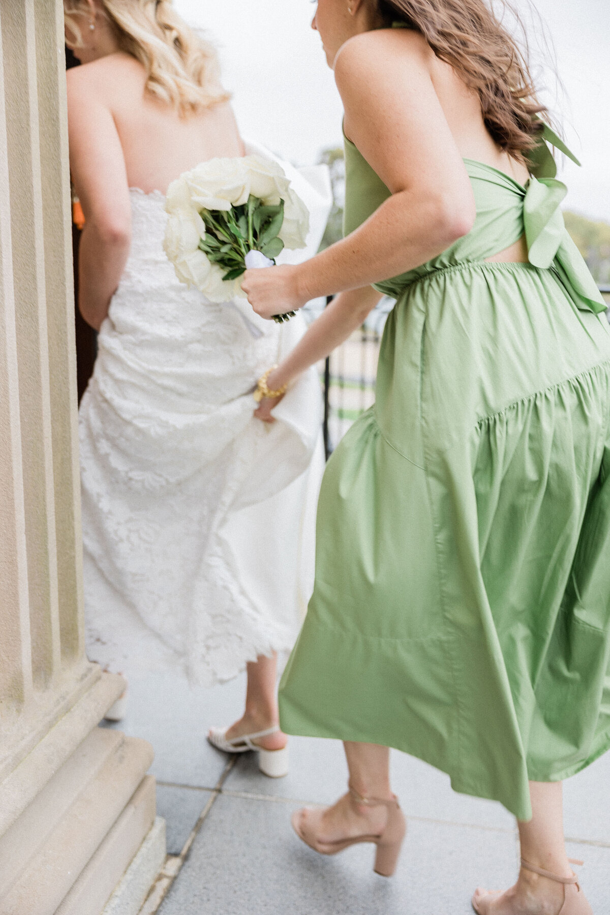 A bridesmaid in a flowing green dress holds the bride’s gown as they ascend stone steps. 
