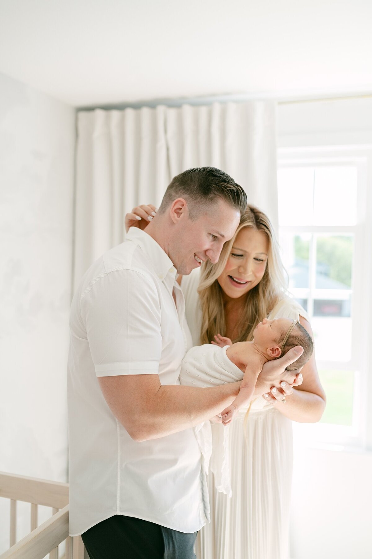 Mom and dad smile at their newborn held in front of them while mom puts her arm around her husband